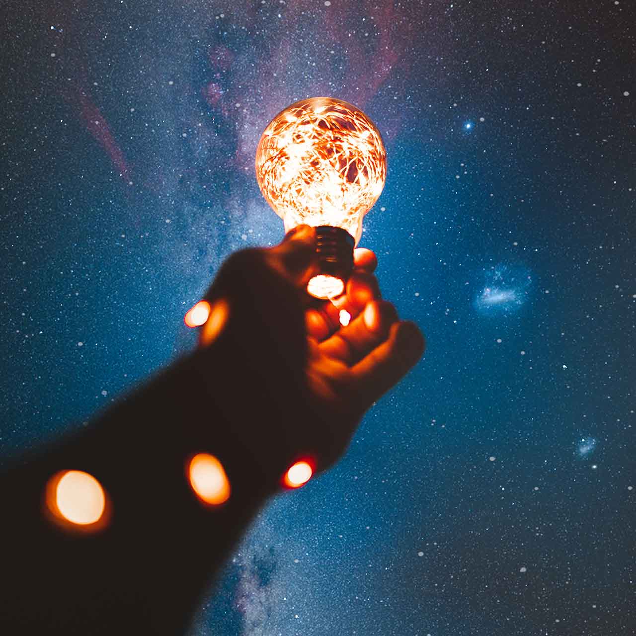 A hand holding a lightbulb with fairy lights up against the night sky with the milkyway visible