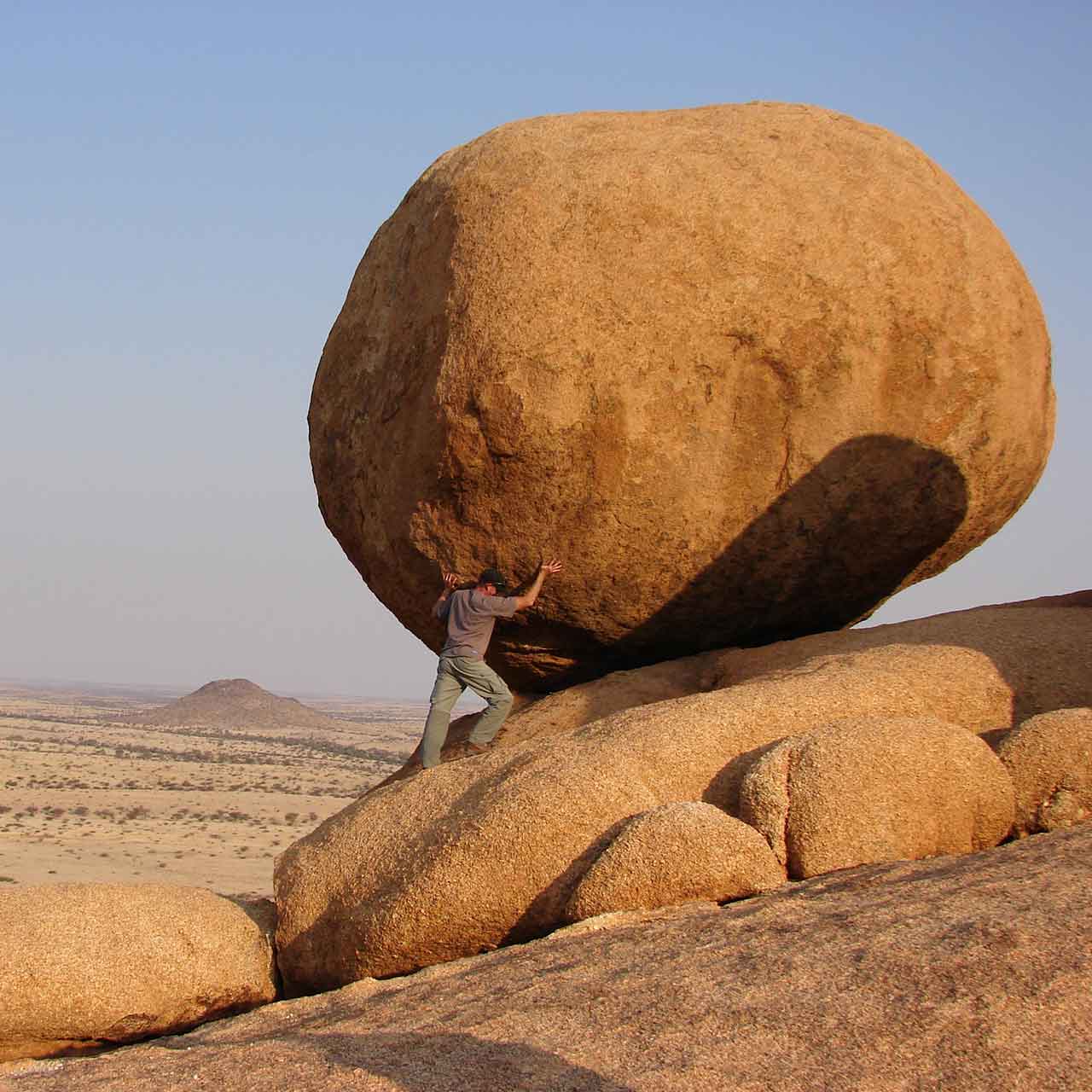 Photo of a man standing below a boulder pretending to lift it